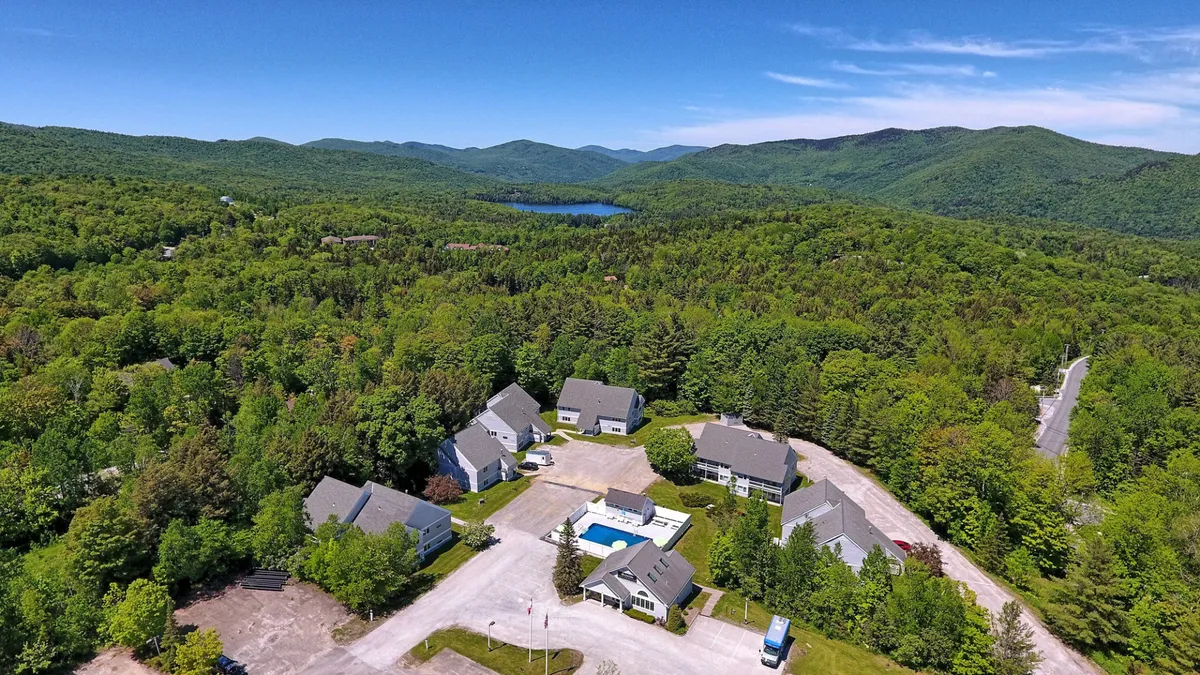 Taken from an exterior vantage, this image presents the wider resort setting with a swimming pool set against a dramatic mountain landscape. You can see a cluster of condo buildings nearby, all nestled among natural greenery. The scene captures the tranquil atmosphere, with elements like morning mist and expansive views, suggesting both privacy and connection to Vermont's beautiful outdoors.