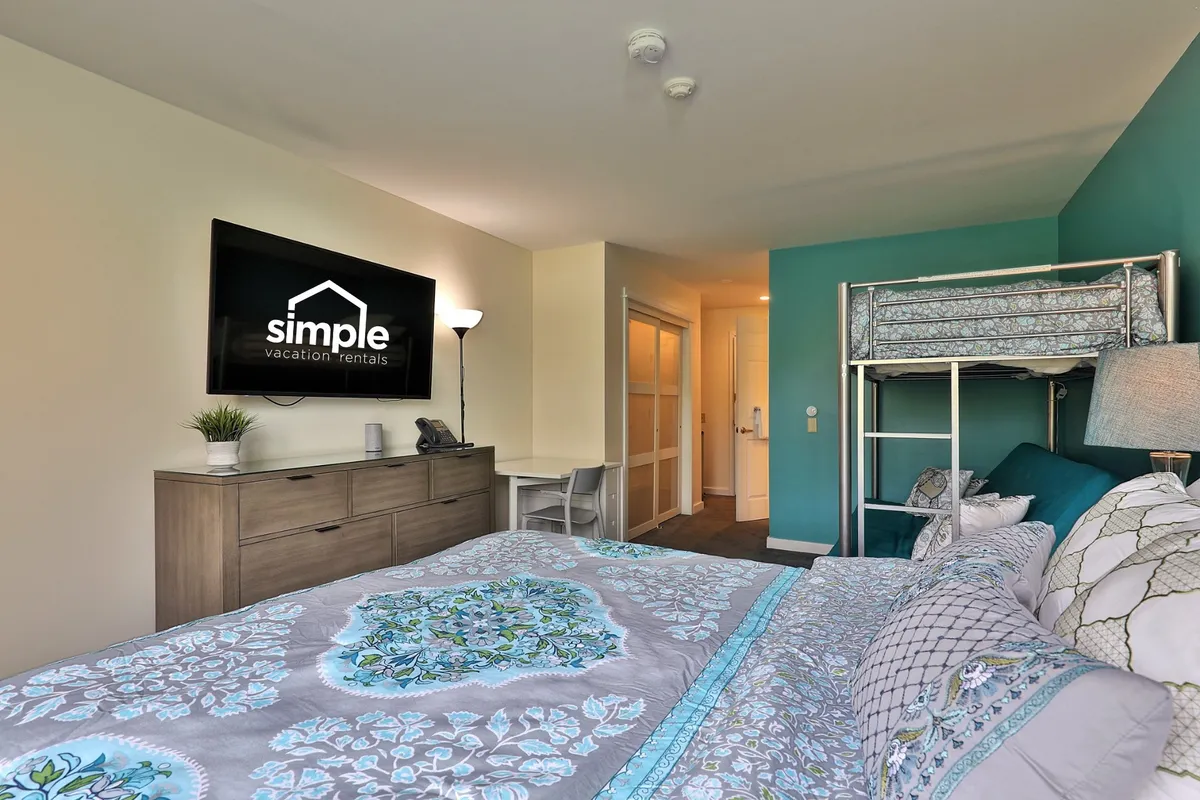 This image spotlights the heart of the main studio area, featuring the plush queen bed draped in a decorative quilt, the adjacent twin-over-full futon bunk, a smart TV positioned for communal viewing, a dresser, and soft lighting from a nearby lamp. The arrangement conveys a harmonious blend of sleeping and lounging zones, ideal for families or groups looking to relax together.