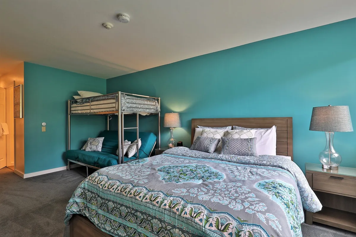 A cozy bedroom featuring both a bunk bed and a main bed, accented with decorative pillows and a nightstand topped with a lamp. The space emanates warmth and comfort, inviting guests to relax, read, or spend quiet time. The room’s arrangement and soft lighting create an ideal nook for winding down or focusing on personal tasks.