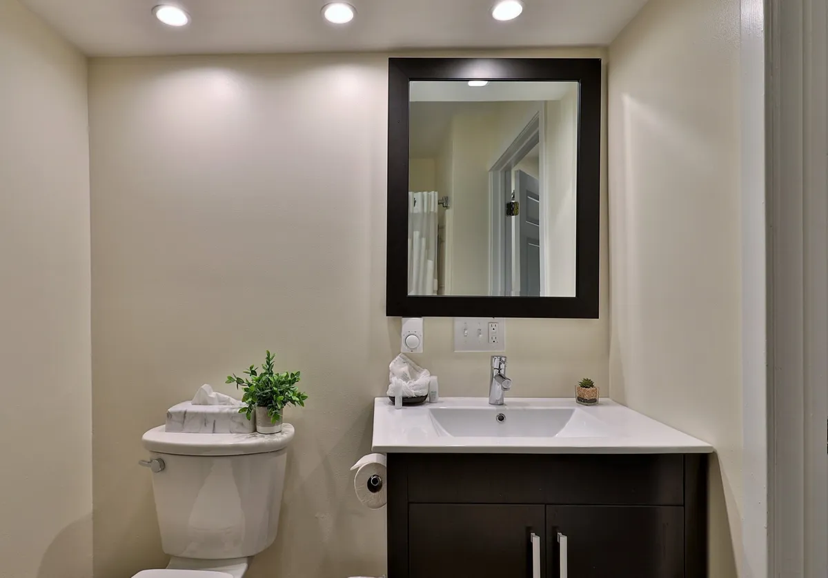A fresh, tidy bathroom scene with a well-lit sink, mirror, toilet, a small potted plant, and a neatly hung towel. The clean lines and calming decor reflect an inviting environment ideal for starting or ending the day with self-care and reflection.