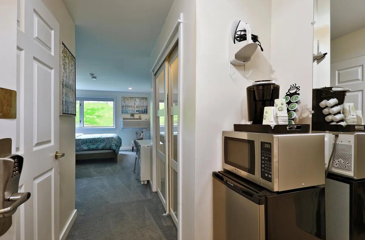 A cozy view into the studio space, highlighting the entryway, compact kitchenette with a mini fridge, microwave, and Keurig coffee maker. The image also shows a small dining nook adjacent to the sleeping area, blending utility with comfort.