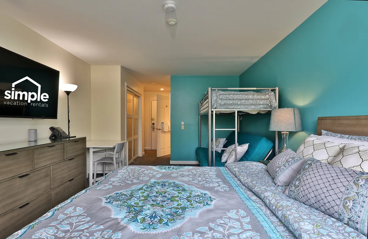 This angle features the bed with crisp decorative bedding, the twin-over-full futon bunk, a wall-mounted TV, and a cheerful blue accent wall. The setup suggests a lively, functional space well-suited for both rest and entertainment, inviting guests to enjoy memorable nights together.