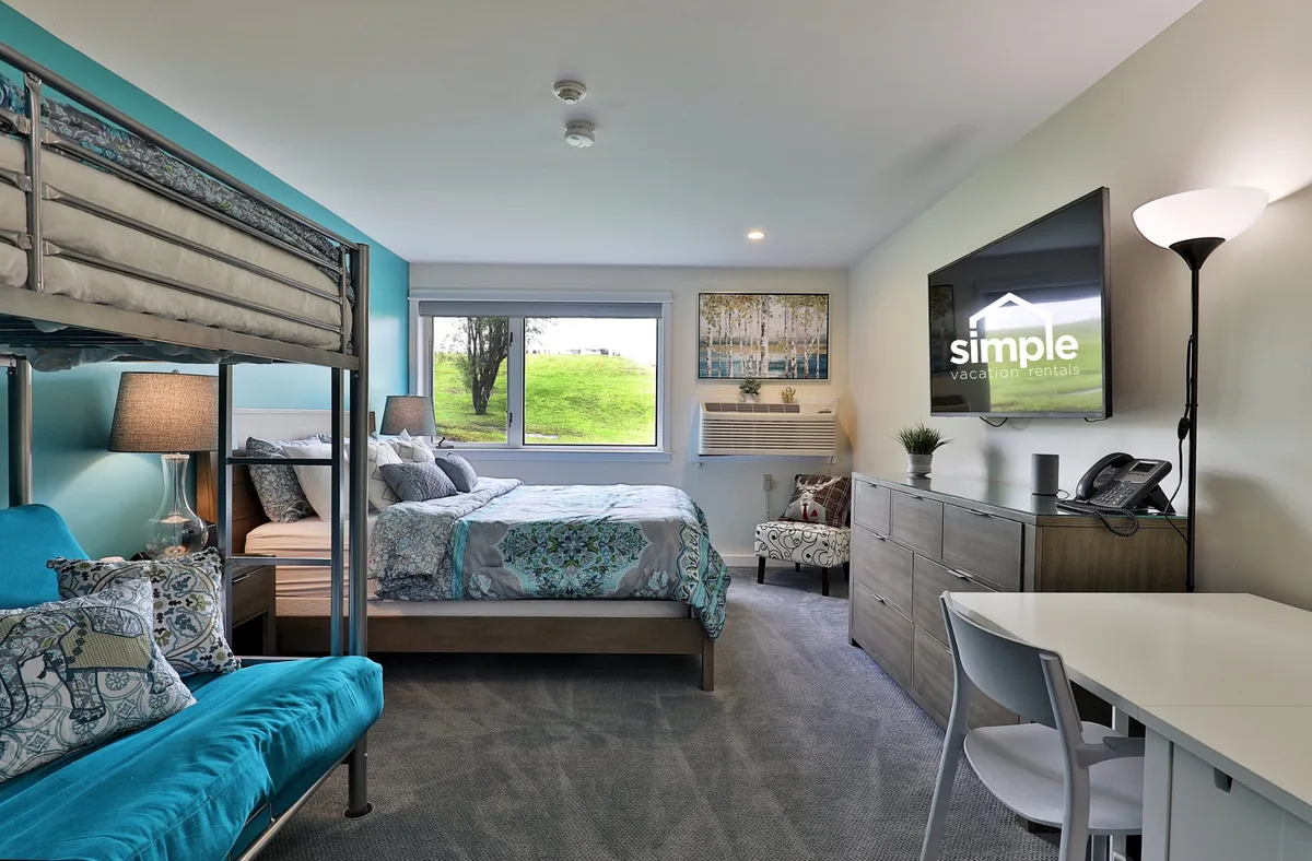 The hero image showcases a bright, inviting studio space with a cozy queen bed and a playful bunk bed, all bathed in natural light from a large window that frames serene Vermont scenery. A modern wall-mounted TV and a versatile dining/work table create a functional yet relaxing environment perfect for unwinding after a day outdoors.