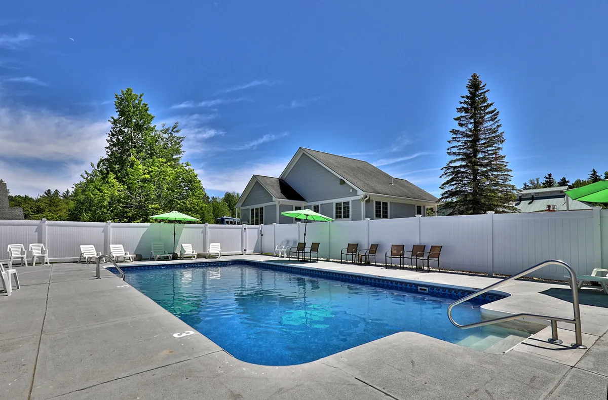 This image showcases the outdoor swimming pool surrounded by white lounge chairs, green umbrellas, and a fenced yard with mature trees. The area is bathed in sunshine, inviting guests to relax poolside, swim, or enjoy a quiet moment with a book. The scene emphasizes leisure, comfort, and the refreshing resort-style amenities available to guests during the warmer months.